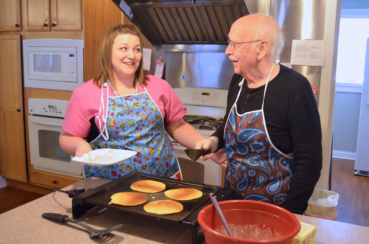 An elderly man and a woman wearing aprons are cooking pancakes on a griddle in a kitchen. The woman is holding a plate and smiling at the man, who is holding a spatula. The kitchen has wooden cabinets, a microwave, an oven, and a stove with a stainless steel hood.