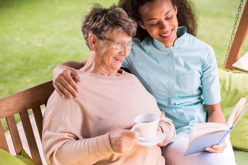 An elderly woman and a caregiver enjoy coffee outdoors on a swing bench.