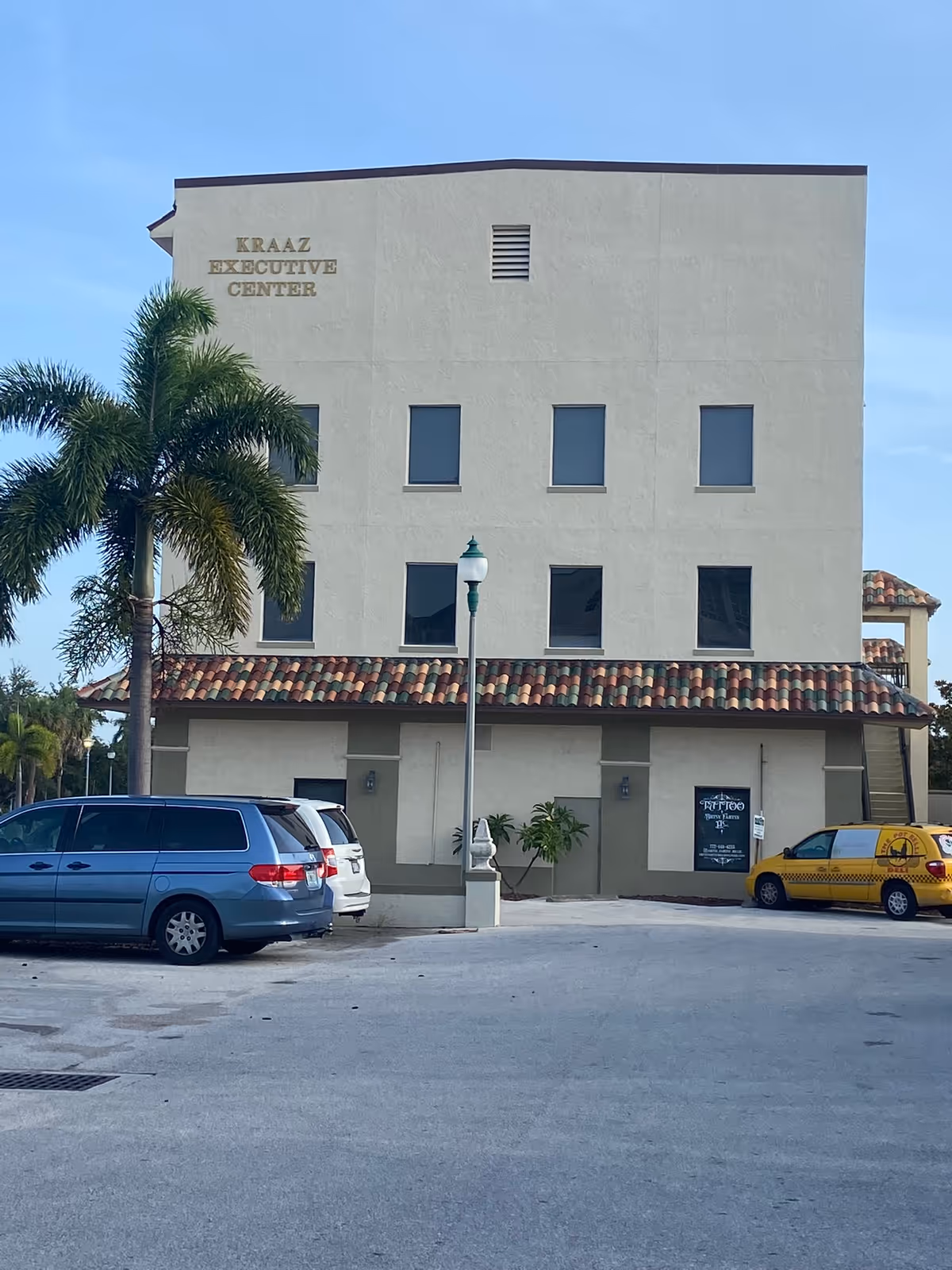 Beige, multi-story building labeled Kraaz Executive Center with palm trees and parked cars in front.