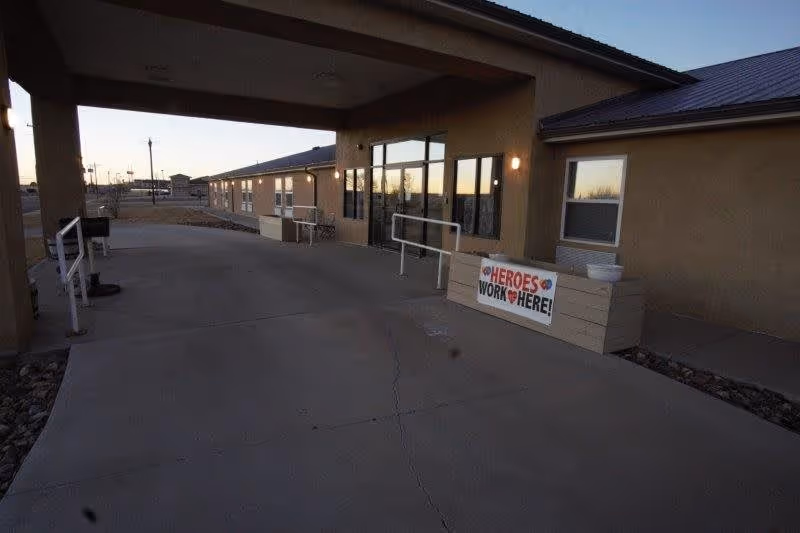 Exterior view of Aspen Leaf Assisted Living Residence in Limon during dusk, showing a covered entrance with a concrete walkway, railings, windows, and a sign that reads 'HEROES WORK HERE!' attached to a planter box.