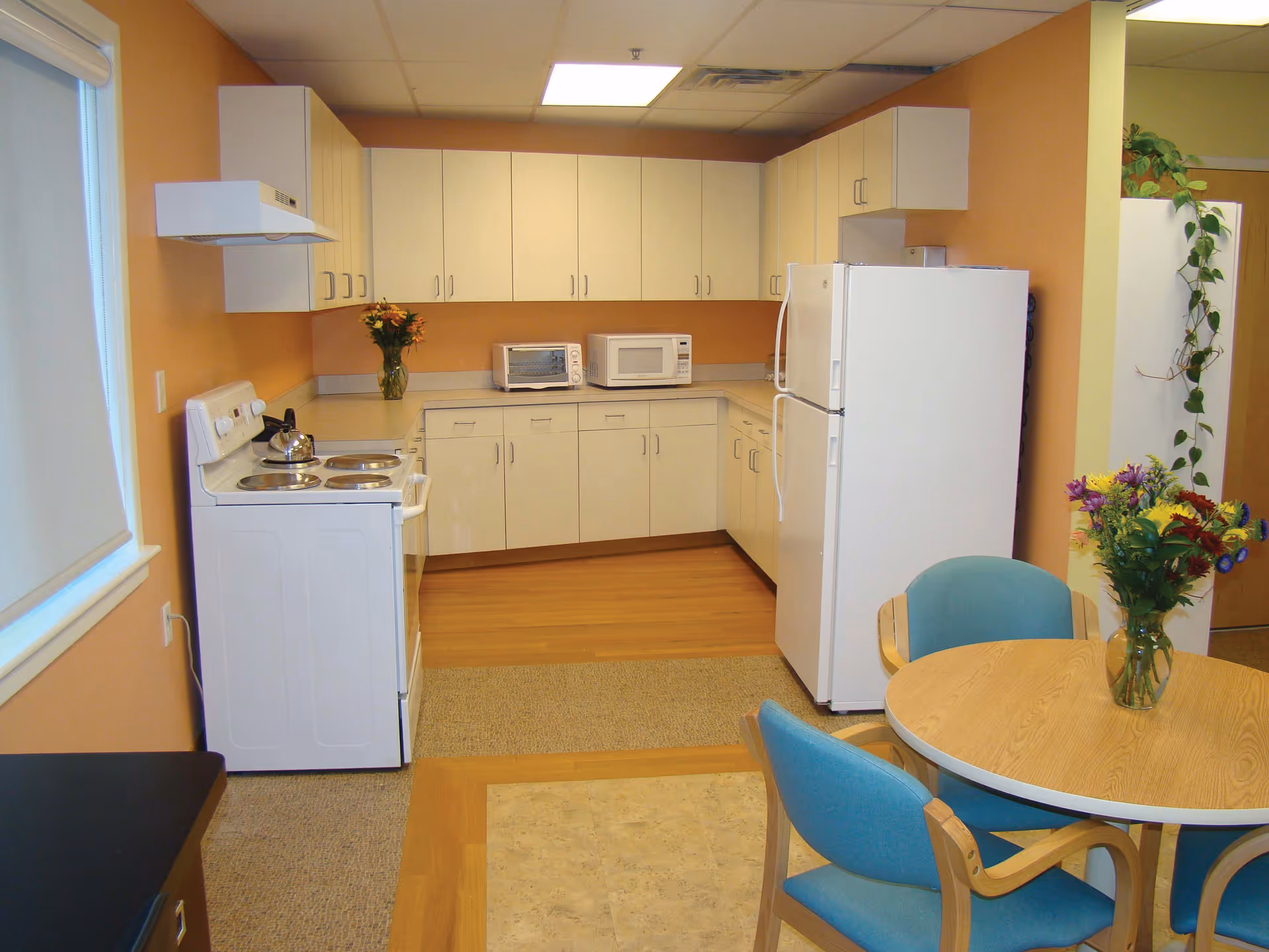A clean kitchen area with white cabinets, a white stove with a kettle, a white refrigerator, a microwave, and a toaster oven. There is a round wooden table with a vase of colorful flowers and three blue cushioned chairs around it. The walls are painted a warm orange color and there is a window with a white blind on the left side.