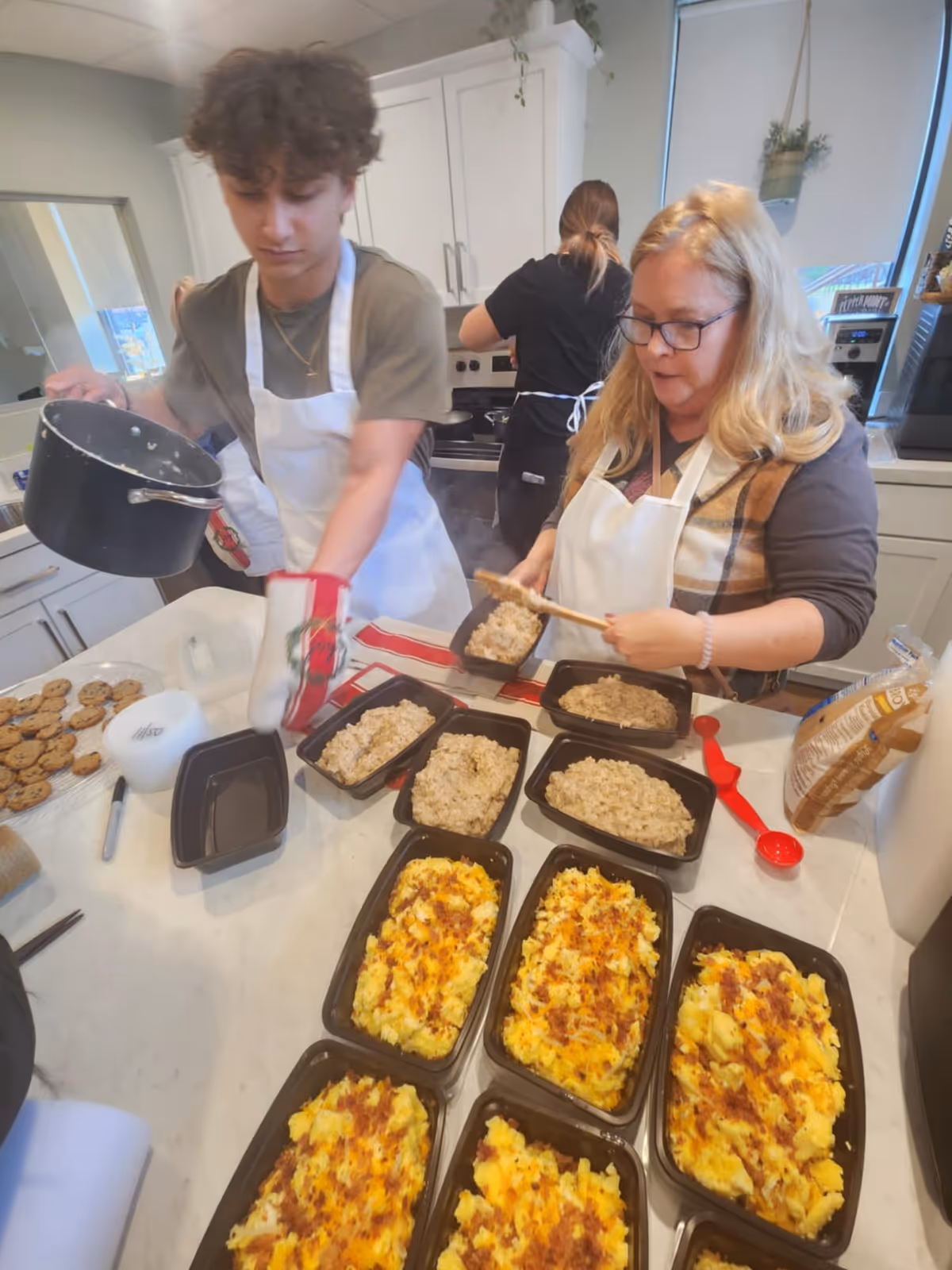 Two staff members prepare meals in a bright kitchen with trays of dishes.