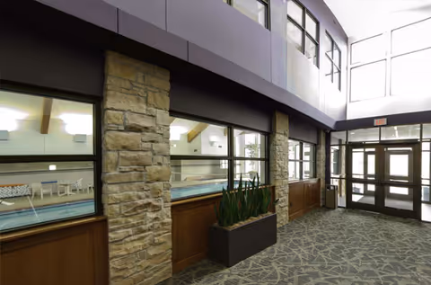 Interior hallway with large windows looking into an indoor swimming pool area. The hallway features stone pillars, a planter with tall green plants, patterned carpet, and glass double doors at the end letting in natural light.