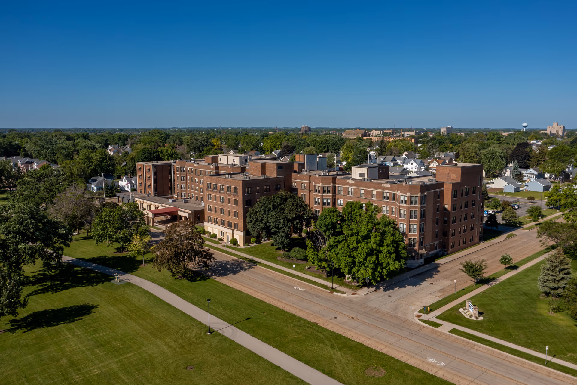 Aerial view of a large multi-story brick residential building with surrounding lawns, trees, streets, and nearby houses under a clear blue sky.