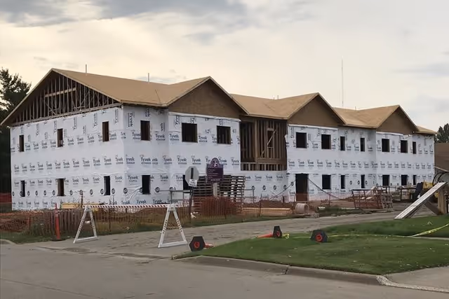 Two-story residential building under construction covered in Tyvek sheathing with construction barriers and equipment in the foreground.