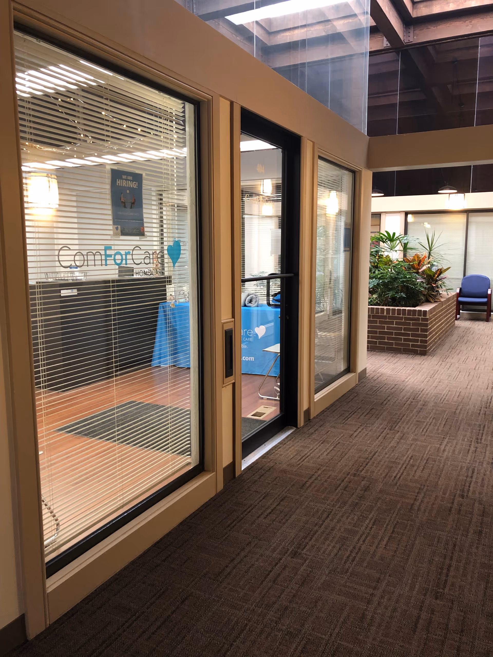 Glass storefronts along a hallway show ComForCare branding, blinds, plants, and seating.