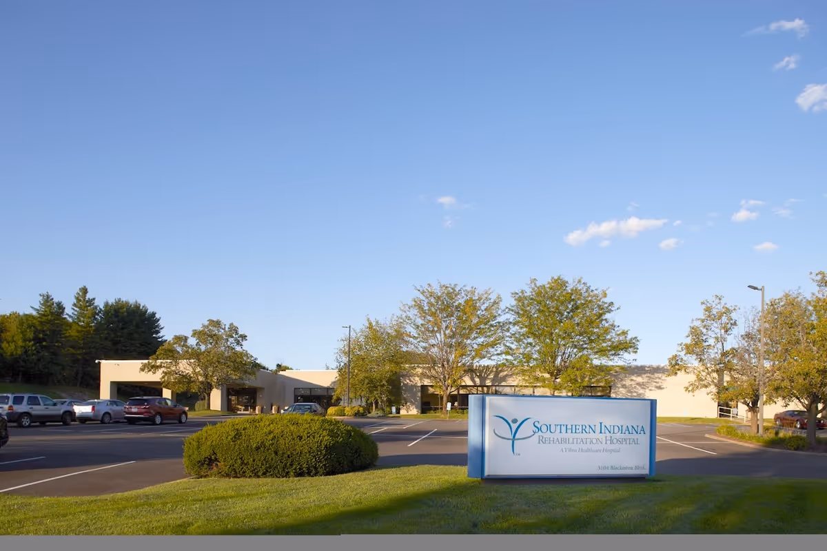 Exterior view of Southern Indiana Rehabilitation Hospital - Skilled Nursing Facility with a parking lot, several cars, trees, and a clear blue sky.