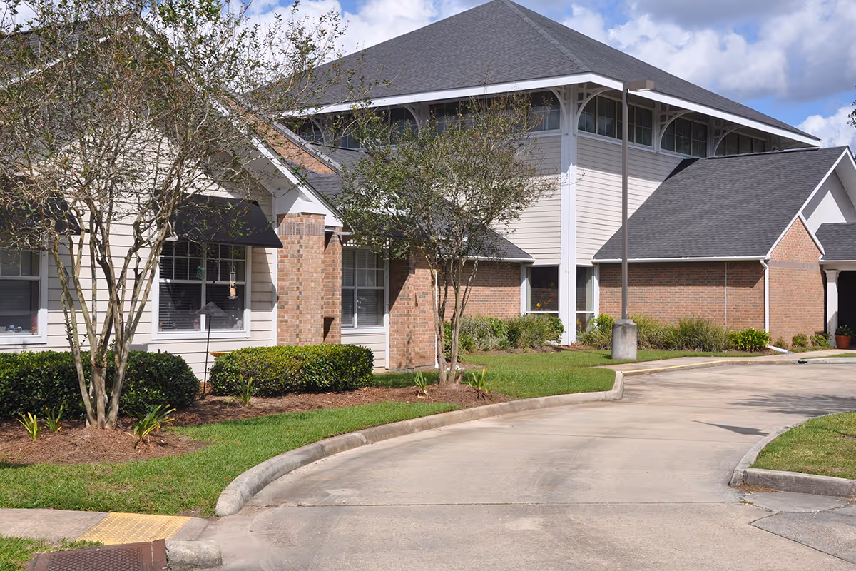 Exterior view of a senior living facility building with beige siding and brick accents, surrounded by trimmed bushes and small trees, under a partly cloudy sky.