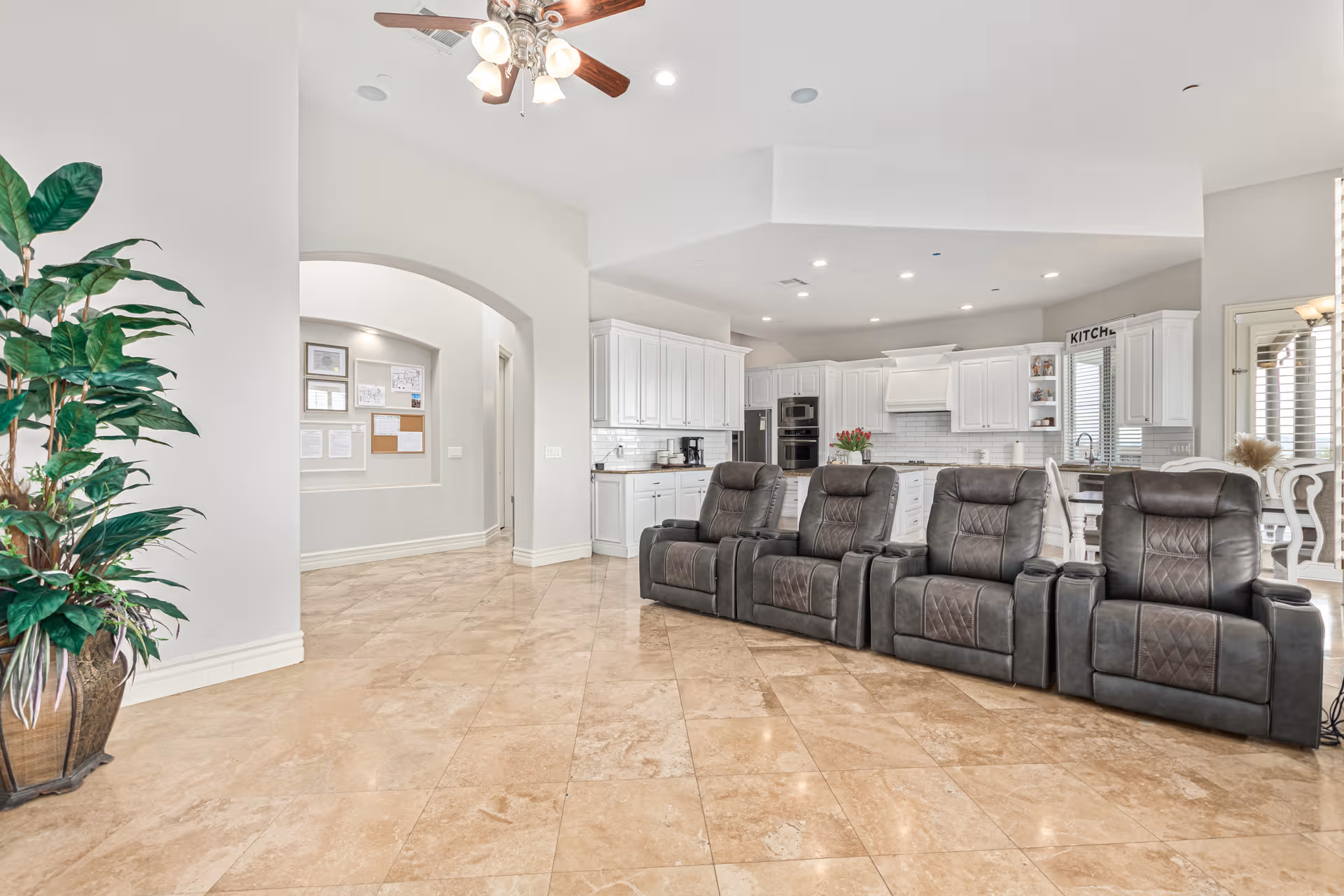 Open living area with gray recliners, white kitchen cabinets, and beige tiled floor.