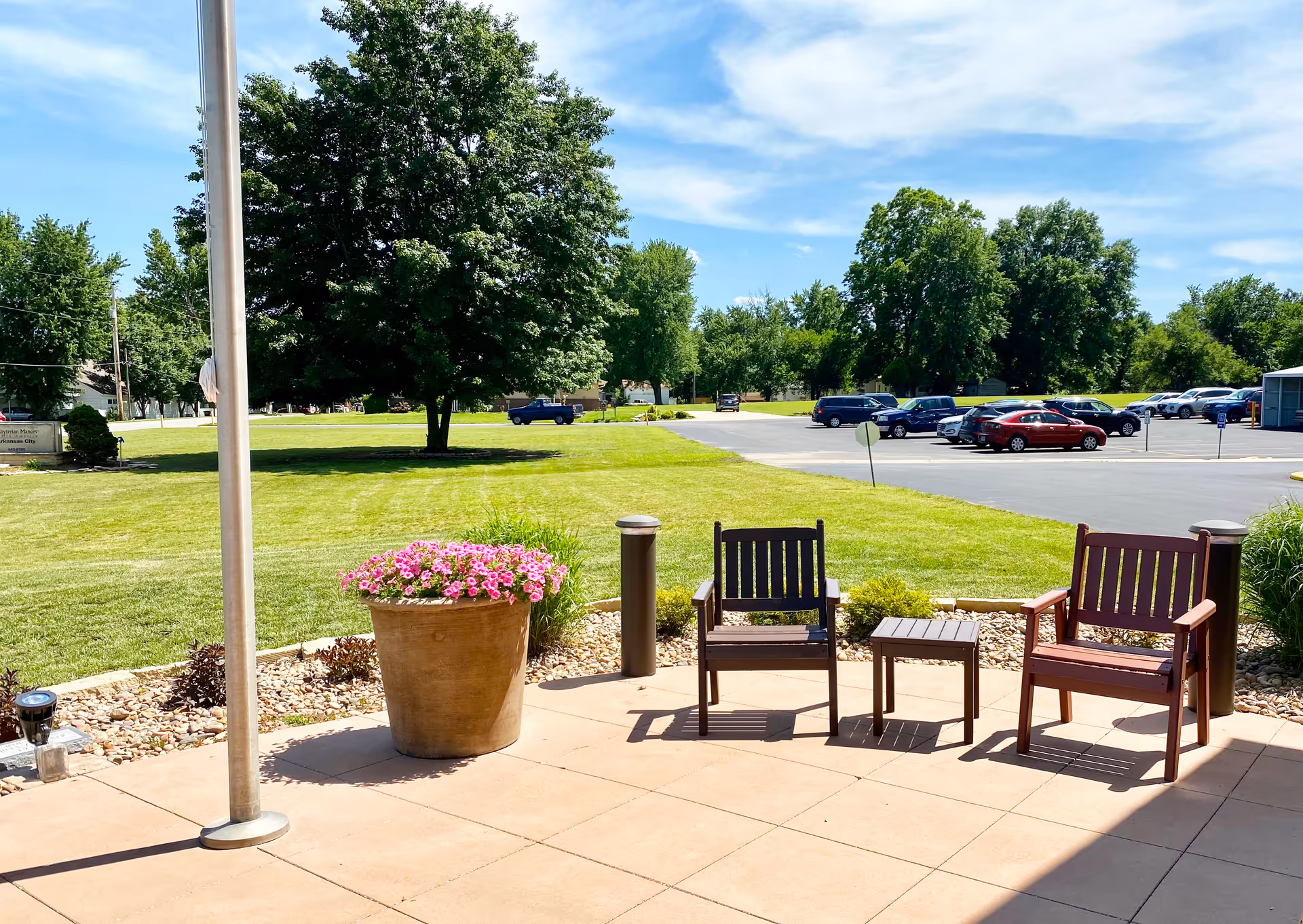 Outdoor patio area with two wooden chairs and a small table between them, a large flower pot with pink flowers, a flagpole, and a view of a green lawn, trees, and a parking lot with several cars under a blue sky with some clouds.