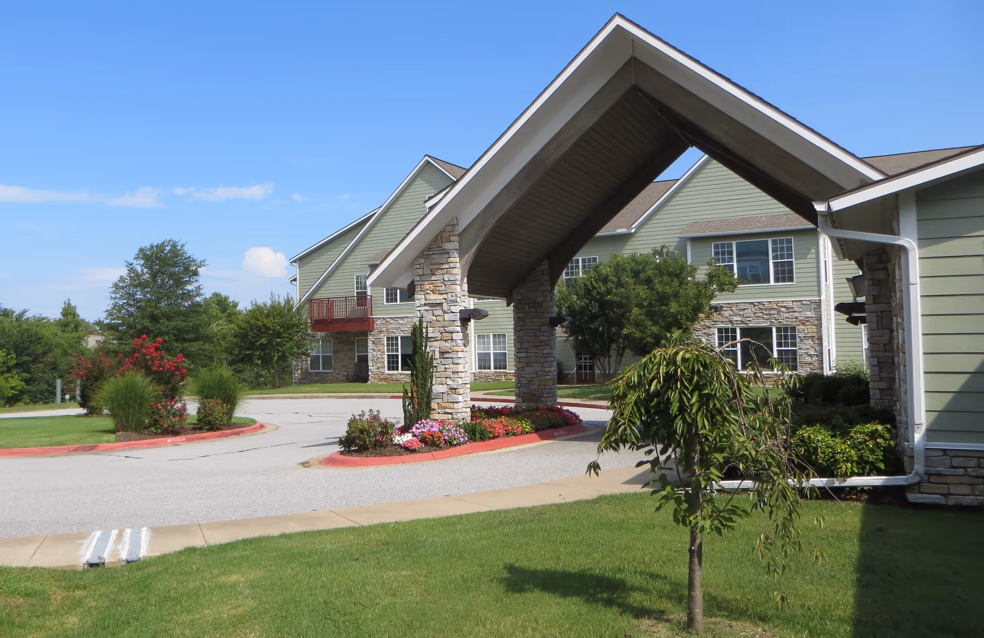 Exterior view of a senior living facility with a covered entrance supported by stone pillars, surrounded by green lawns, flowering plants, and trees under a clear blue sky.