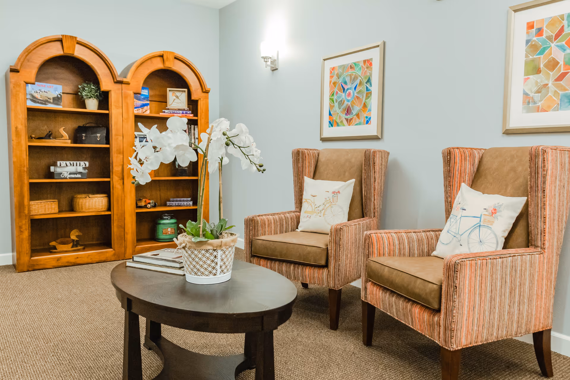 A cozy sitting area with two striped armchairs featuring brown cushions and decorative pillows with bicycle designs. Between the chairs is a dark wooden oval coffee table with a white potted orchid plant and some books. Behind the chairs, there are two colorful framed artworks on a light blue wall. To the left, there is a wooden bookshelf with various decorative items and books.