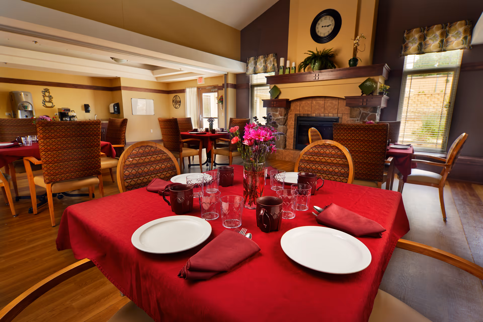 A dining room with tables covered in red tablecloths, set with white plates, maroon napkins, glasses, and maroon mugs. There is a vase with pink flowers on the table in the foreground. The room has wooden floors, patterned chairs, a fireplace with a clock above it, and windows with patterned valances letting in natural light.