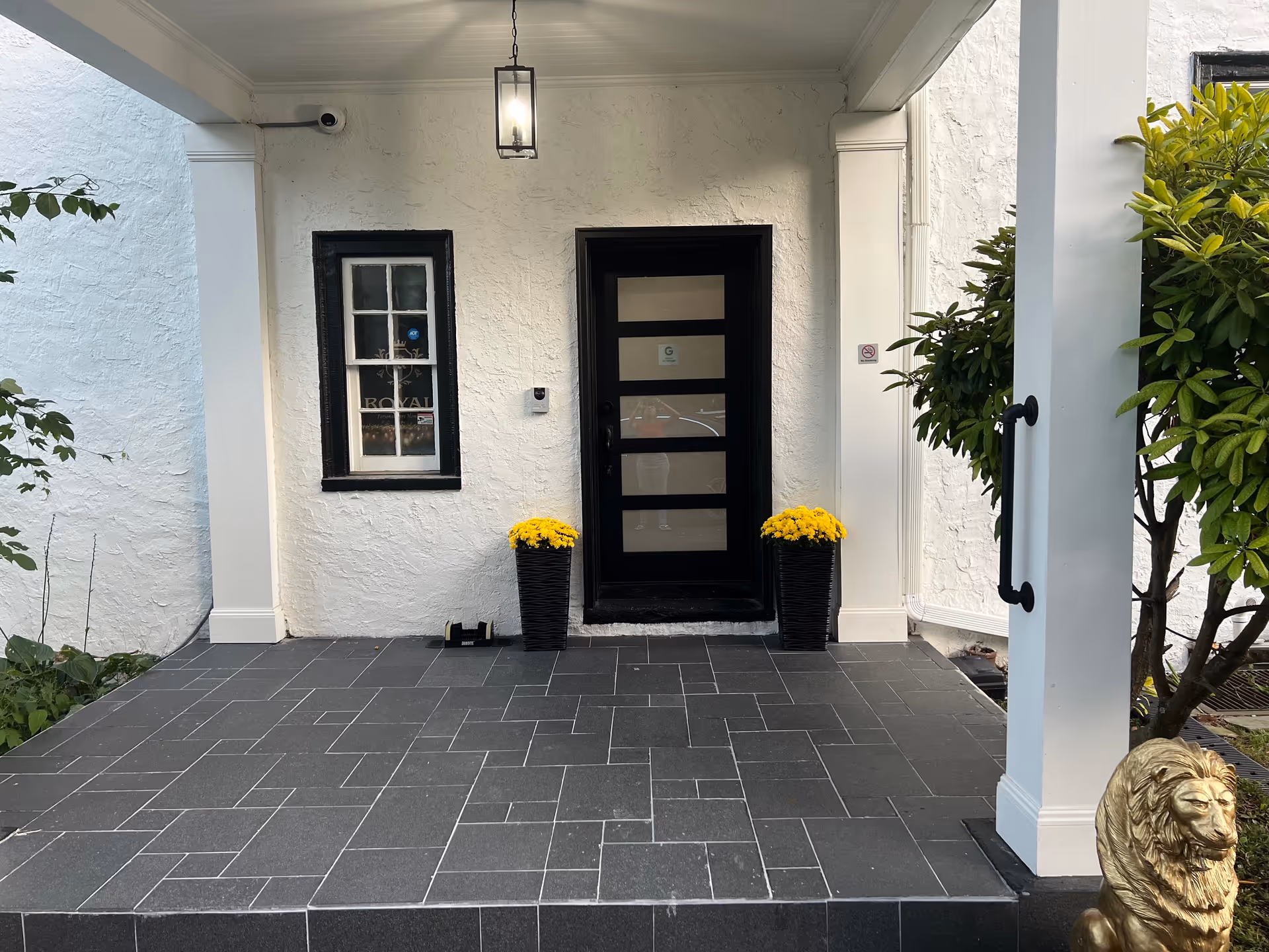 White textured exterior entry with black front door, yellow potted flowers, and tiled porch.