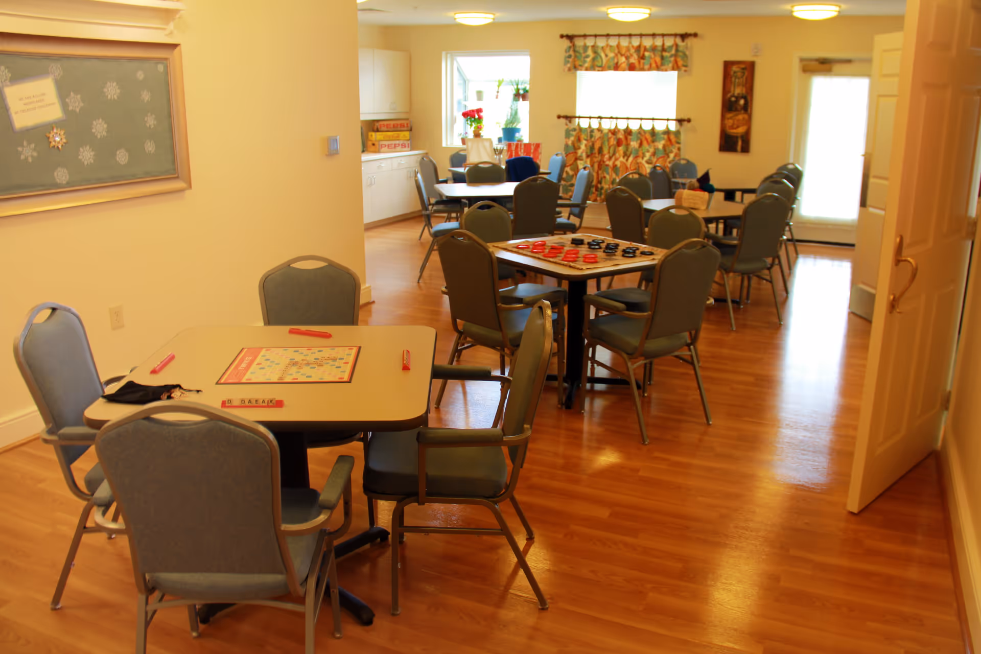 A well-lit room with several tables and chairs arranged for group activities. One table has a Scrabble board game set up, and another table has a checkers board. The room has wooden flooring, light-colored walls, a bulletin board with snowflake decorations, and windows with colorful curtains letting in natural light.