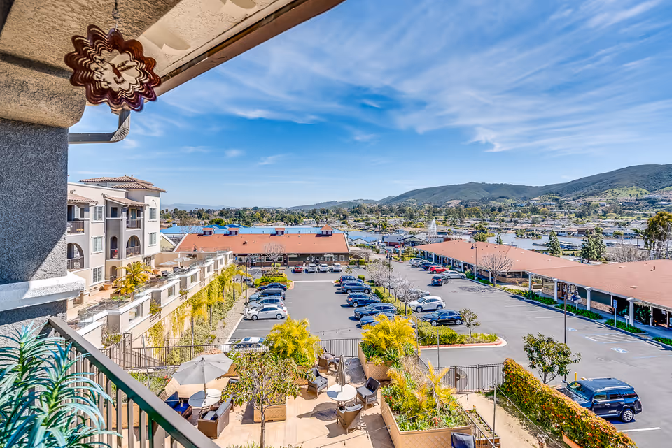 Balcony view over a courtyard and parking lot toward red-roofed buildings and distant hills under a blue sky.