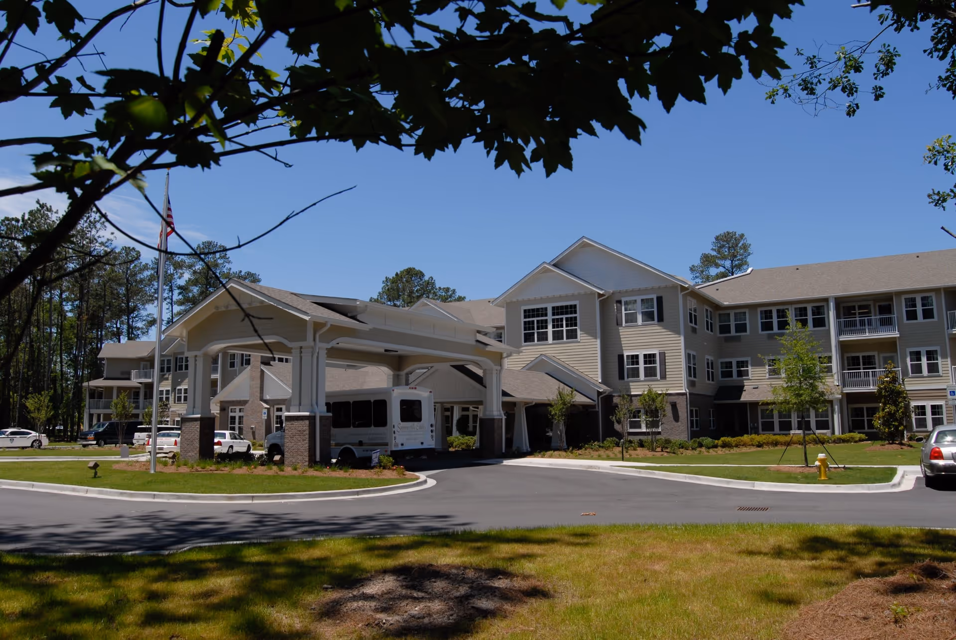 Exterior view of Summerville Estates, a multi-story senior living facility with beige siding and brick accents. The building features a covered entrance with a driveway where a white shuttle bus is parked. There are several cars parked nearby, a flagpole with an American flag, and trees surrounding the property under a clear blue sky.