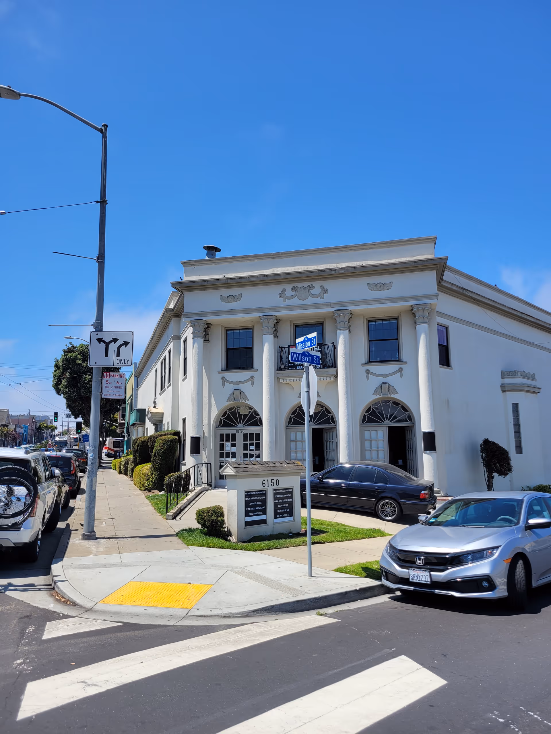 White neoclassical building on a street corner with arched windows and parked cars.