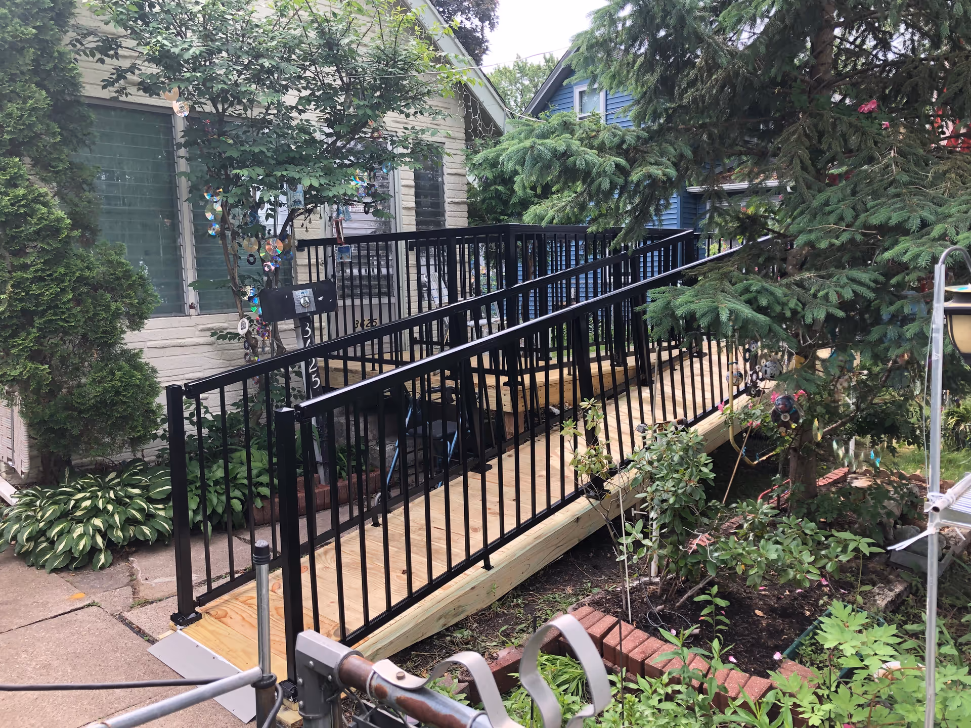 Wooden ramp with black railing leads to a house entrance amid shrubs and trees.