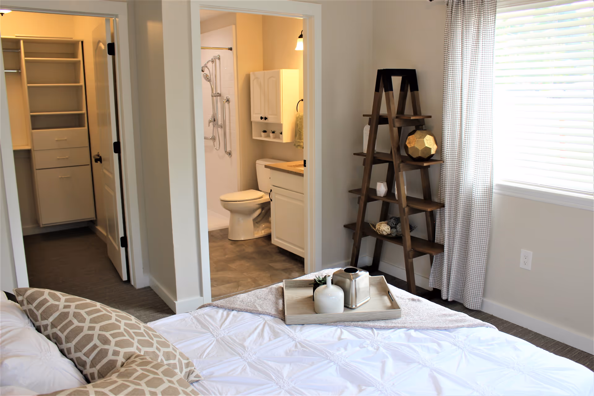 A bright bedroom with a neatly made bed featuring patterned pillows and a tray with decorative items. To the right, there is a wooden ladder shelf with various decor pieces. In the background, an open door reveals a bathroom with a toilet, vanity, and shower equipped with grab bars. Another open door leads to a walk-in closet with shelves and drawers. A window with white blinds and checkered curtains allows natural light into the room.