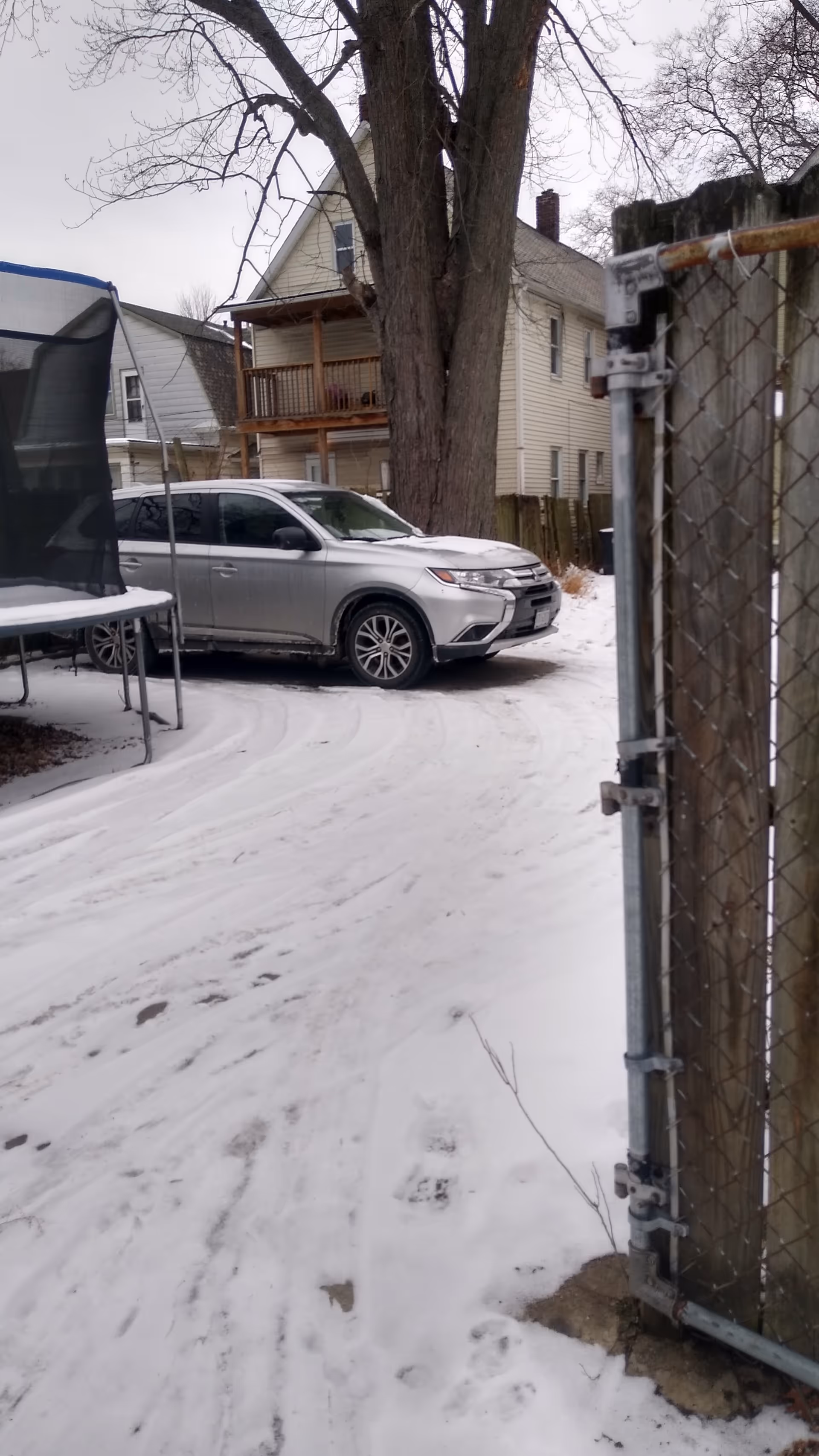 Snow-covered driveway with a parked car, trampoline, and chain-link fence near a house.