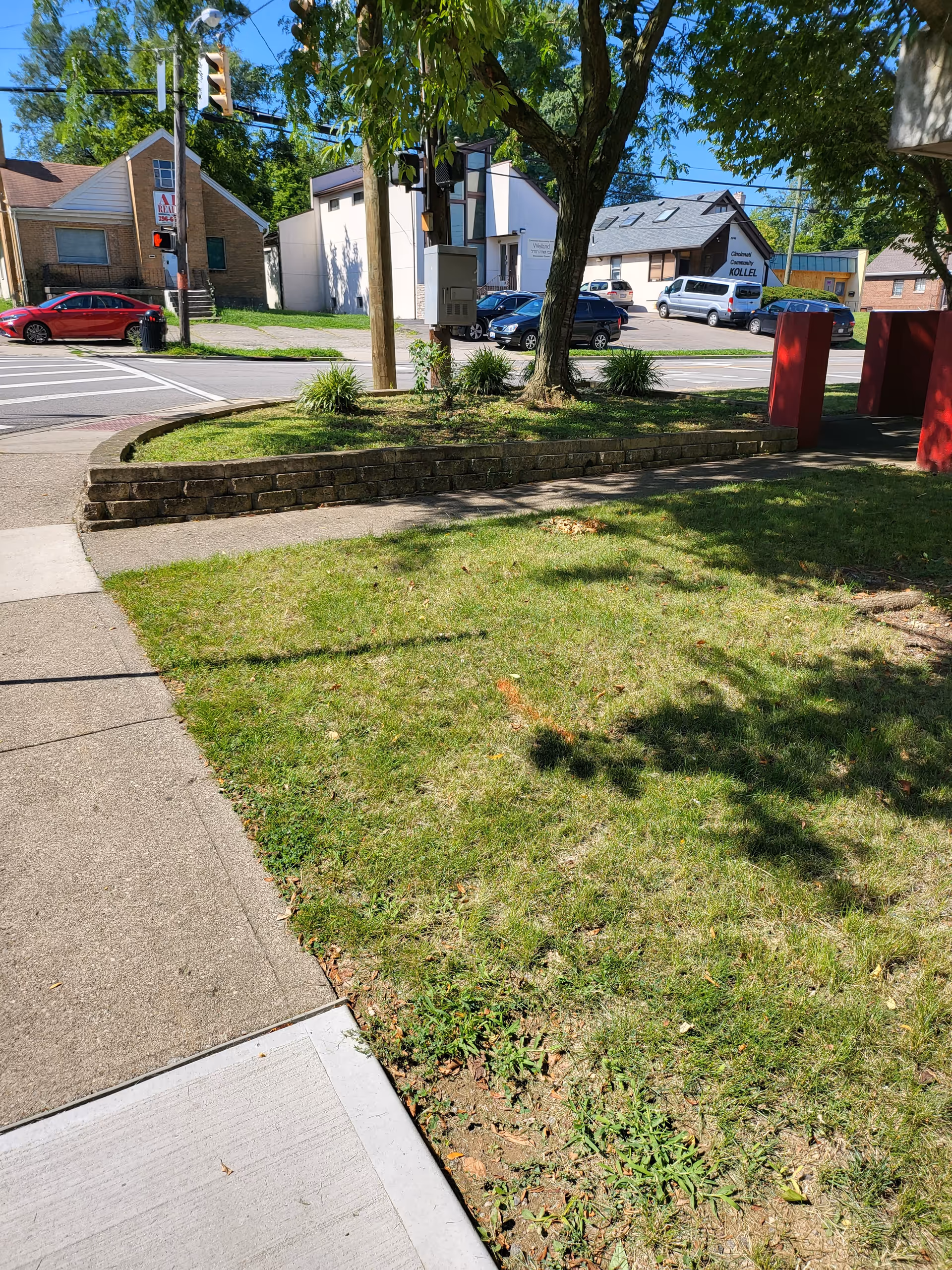 Exterior street view with grassy yard, brick planter, trees, and parked cars along the curb.
