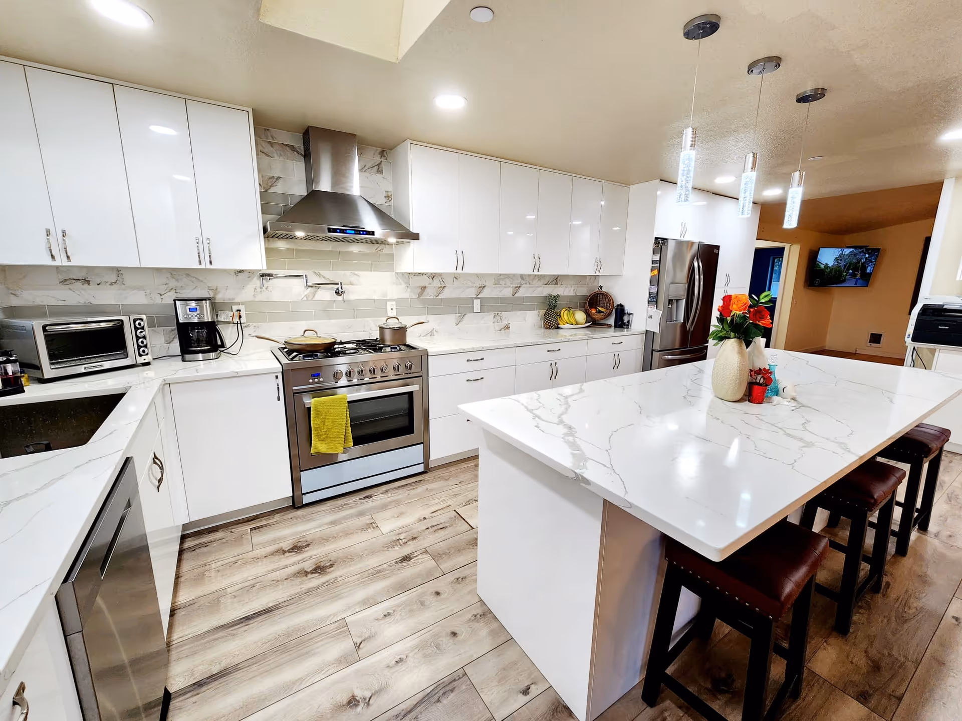 Modern kitchen with white cabinets, marble countertops, stainless steel appliances including a stove, oven, refrigerator, toaster oven, and coffee maker. There is a large kitchen island with a vase of flowers and three brown stools underneath. The floor is wood with a light finish and the ceiling has recessed lighting and three pendant lights above the island.