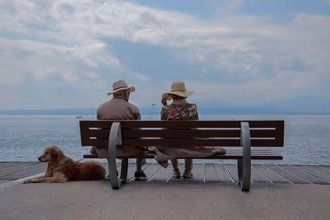 Two elderly people sit on a bench by a lake, with a dog resting beside them.