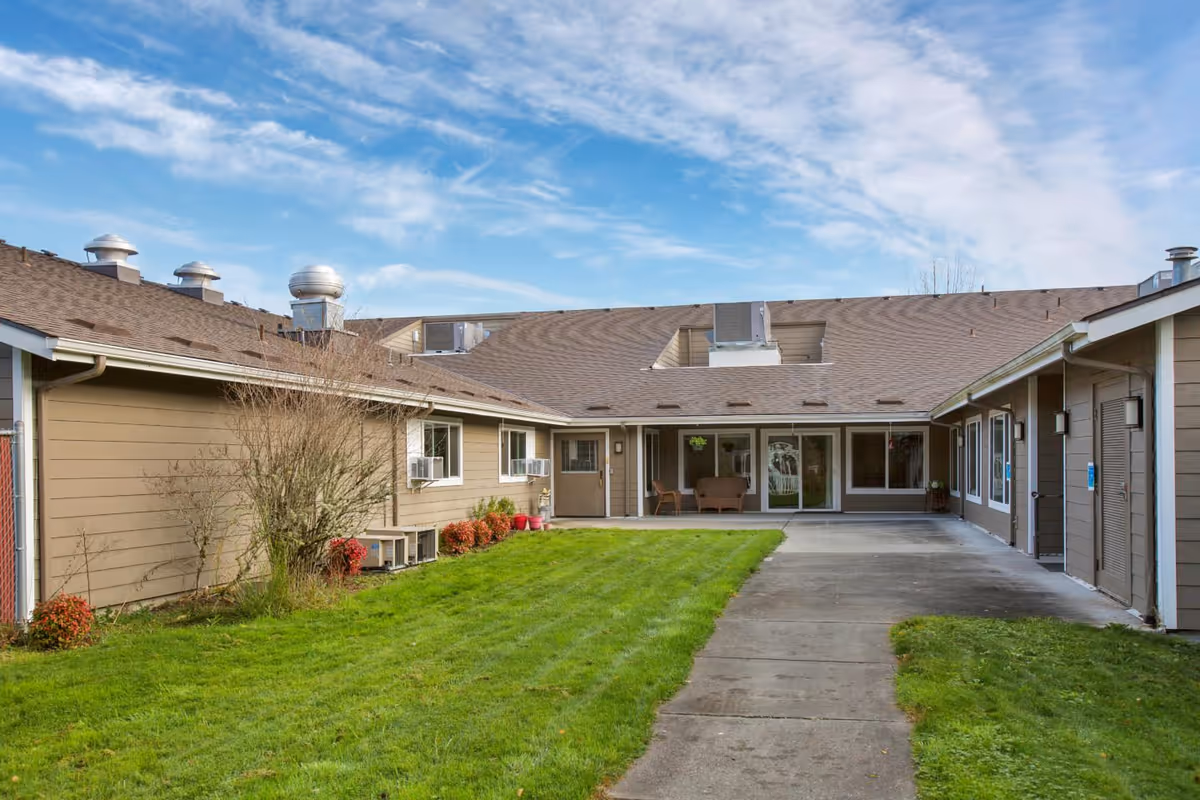 Courtyard of a single-story rehab facility with a grassy lawn, concrete walkway, and a covered patio with seating.