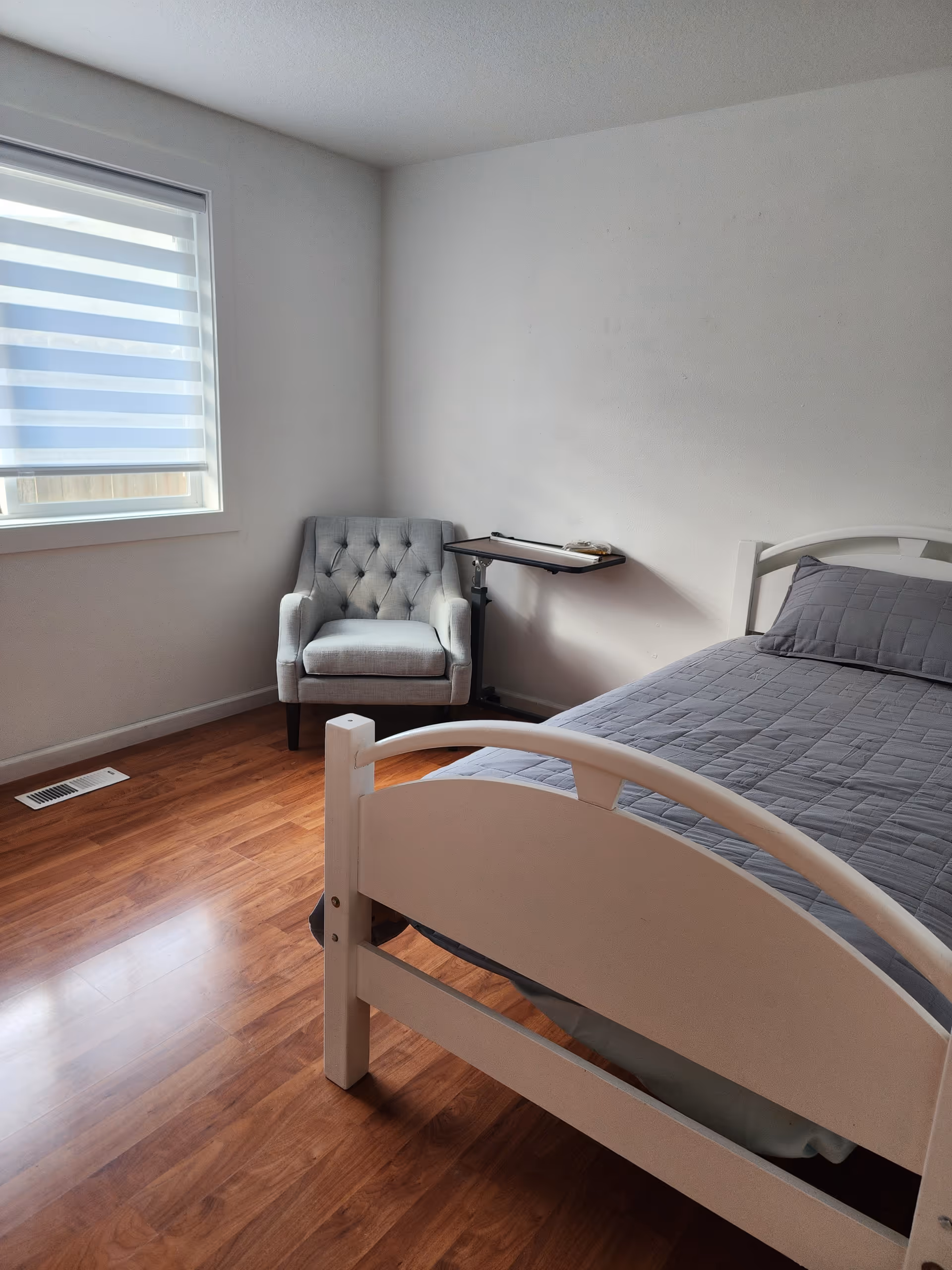 Bright bedroom in a senior living facility with a bed, grey quilt, chair, and window blinds.