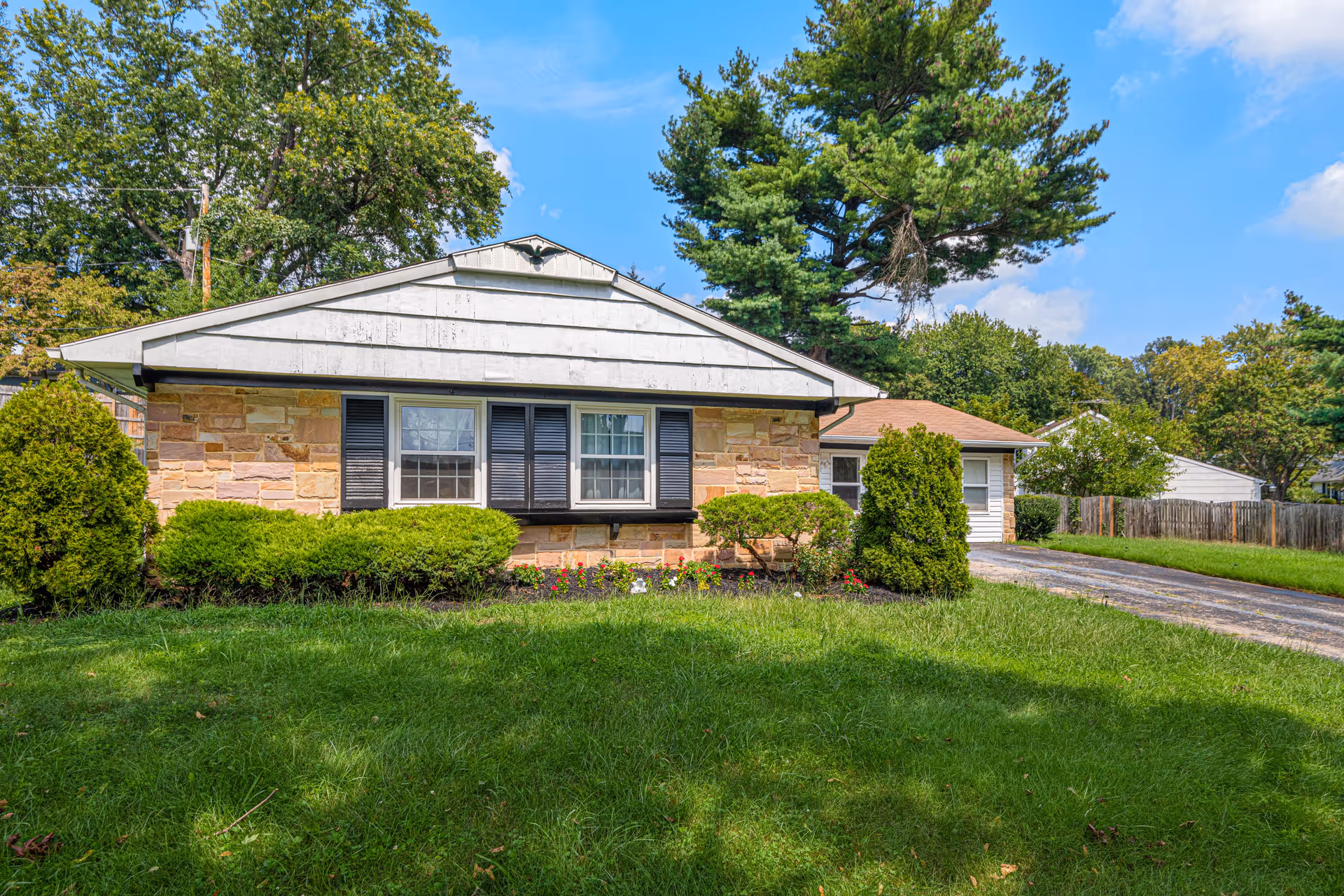 Front exterior view of a single-story residential building with stone and white siding, black shutters on windows, surrounded by green bushes and a well-maintained lawn under a blue sky with some clouds.
