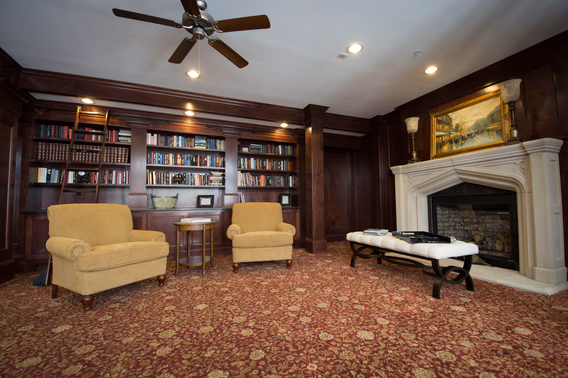A cozy library or lounge area with two beige armchairs, a small round wooden table between them, a large built-in wooden bookshelf filled with books, a ceiling fan, and a white stone fireplace with a painting above it. The floor is covered with a red patterned carpet.