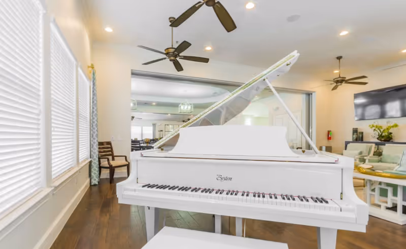 White grand piano in a bright senior living lounge with ceiling fans and seating.