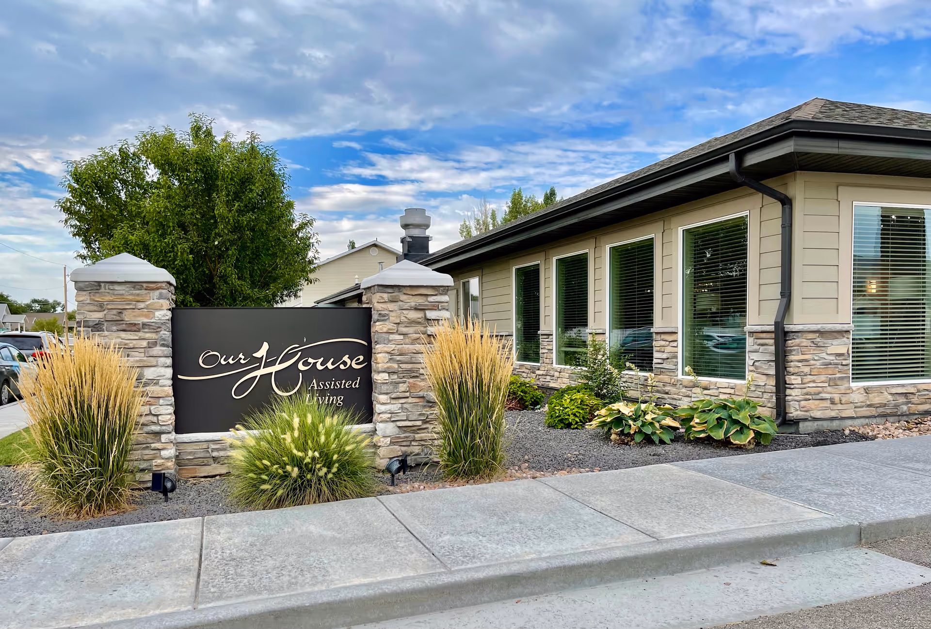 Exterior view of Our House Assisted Living of Tremonton showing a stone and metal sign with the facility name, surrounded by ornamental grasses and plants. The building has beige siding with stone accents and multiple windows with blinds. The sky is partly cloudy.