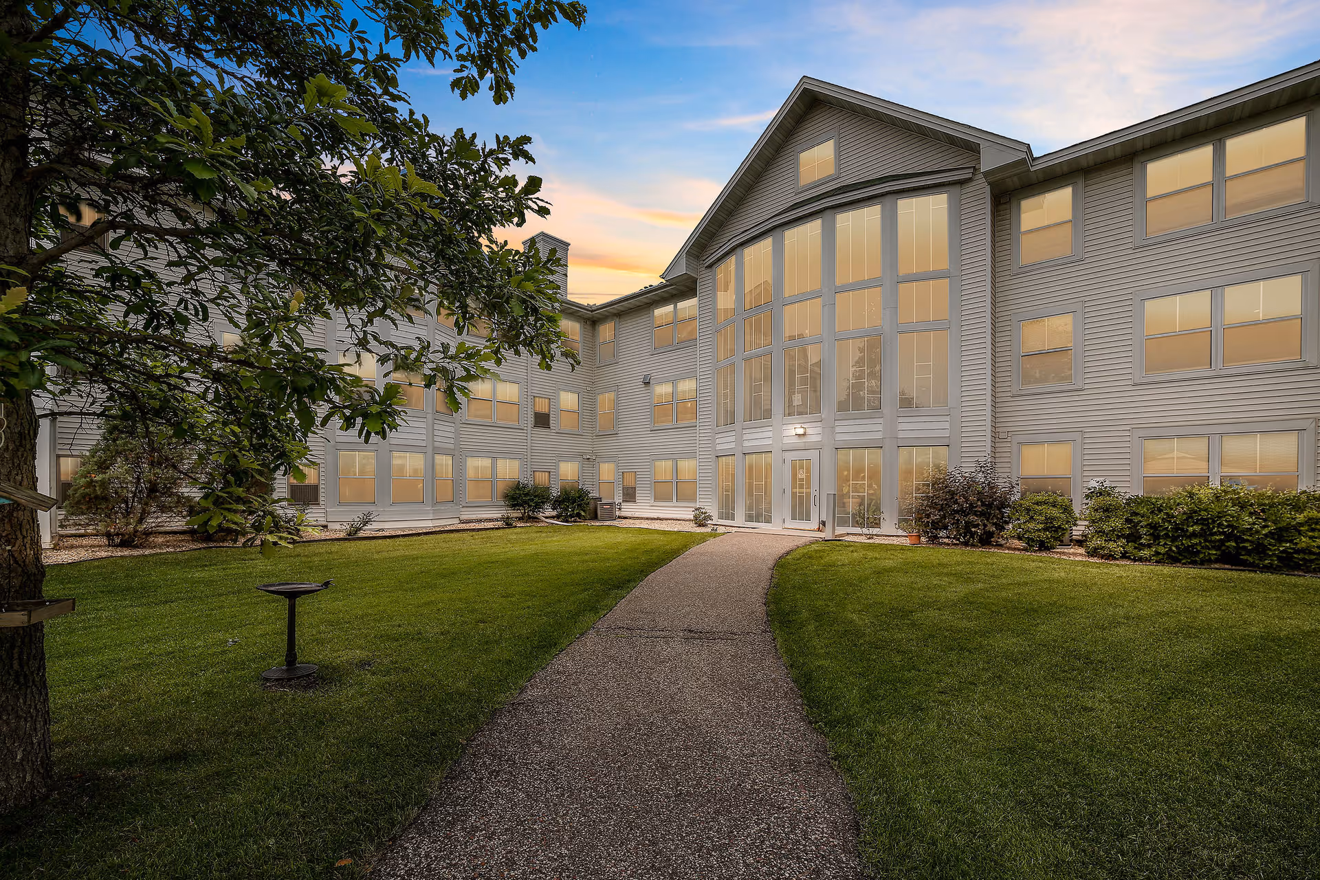 Exterior view of a three-story senior living facility building at sunset with large windows and a central glass entrance. A paved pathway leads through a well-maintained green lawn with bushes and a tree on the left side.