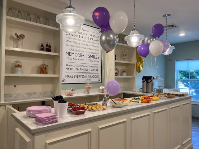 A buffet-style counter in a dining area decorated with purple, white, and silver balloons. The counter has plates, napkins, utensils, and various food items including fruits and snacks. Behind the counter are shelves with decorative items and a framed sign with an inspirational quote about sweetness and memories. Pendant lights hang from the ceiling, and a window with a view of greenery is visible in the background.