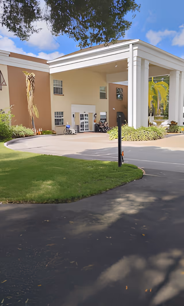 Front entrance of a tan senior living building with a covered porte-cochère, white columns, and a landscaped driveway.