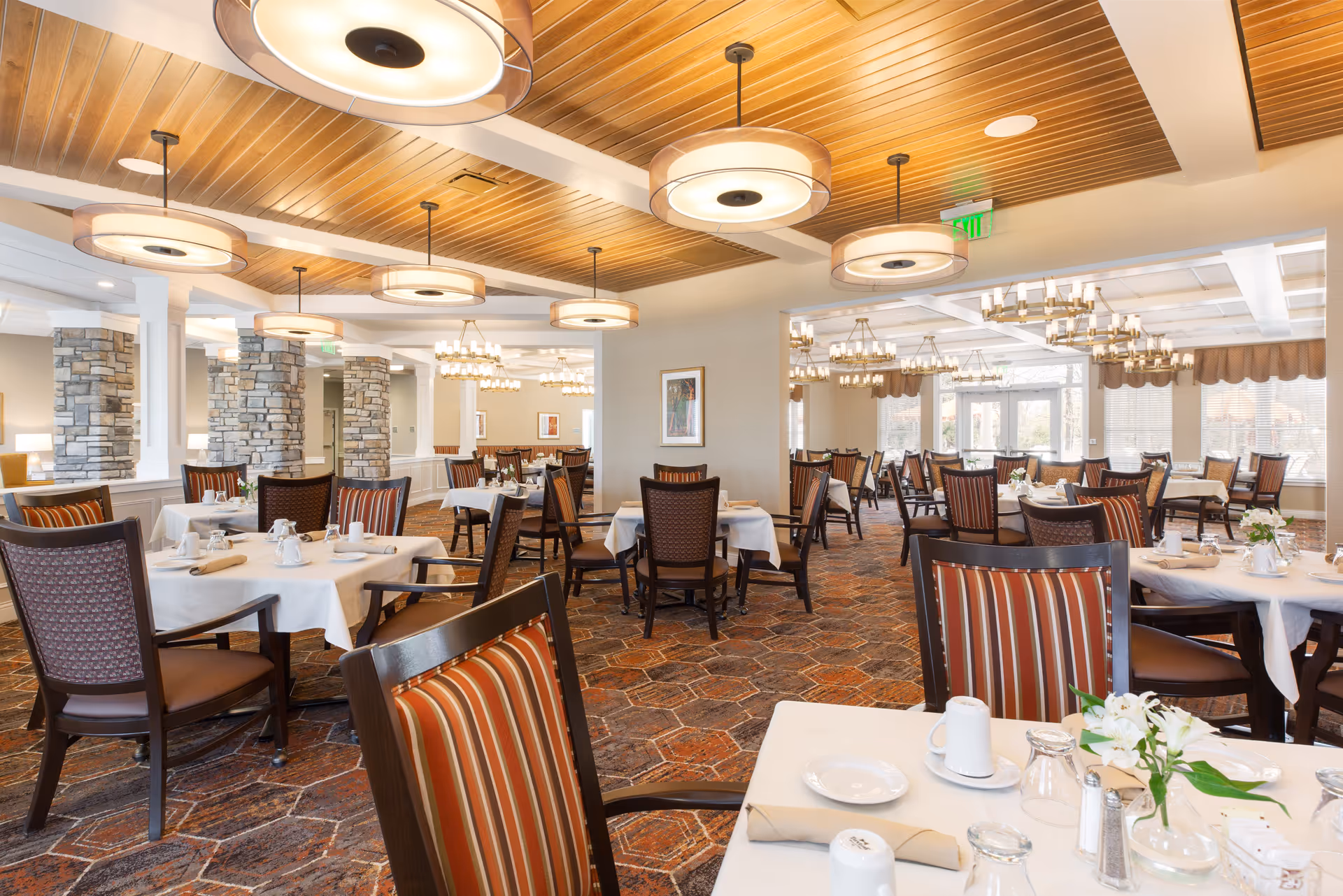 A spacious dining room with multiple tables covered in white tablecloths, set with plates, cups, and napkins. The room features wooden chairs with striped upholstery, a patterned carpet, stone pillars, and modern circular ceiling lights. Large windows allow natural light to fill the space.