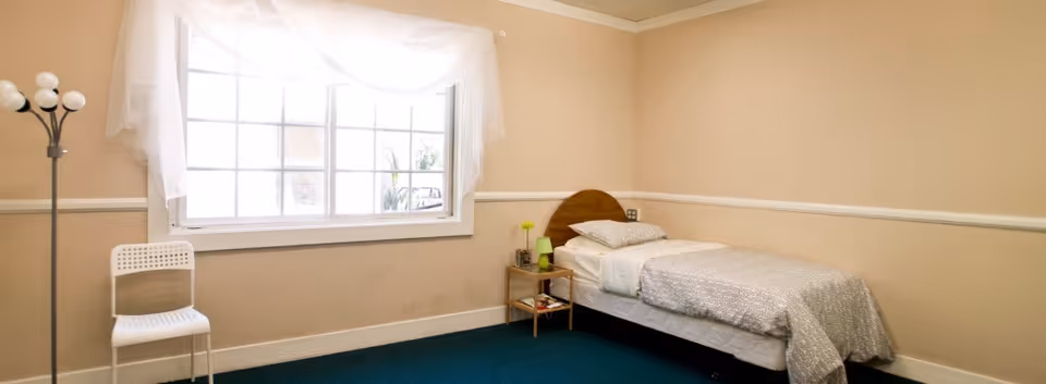 A simple bedroom with a single bed against the right wall, covered with white and gray bedding. Next to the bed is a small wooden nightstand with a green lamp and a small vase with a yellow flower. A large window with sheer white curtains is on the left wall, letting in natural light. A white chair and a floor lamp with multiple white bulbs are positioned near the window. The walls are beige with white trim, and the floor is covered with dark green carpet.