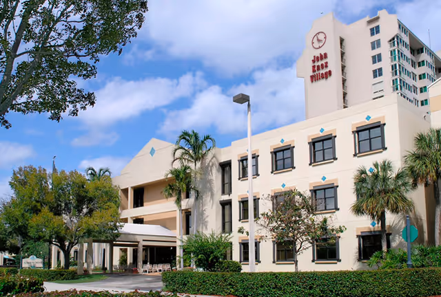 Three-story senior living facility with tan walls, palm trees, and a John Knox Village sign.