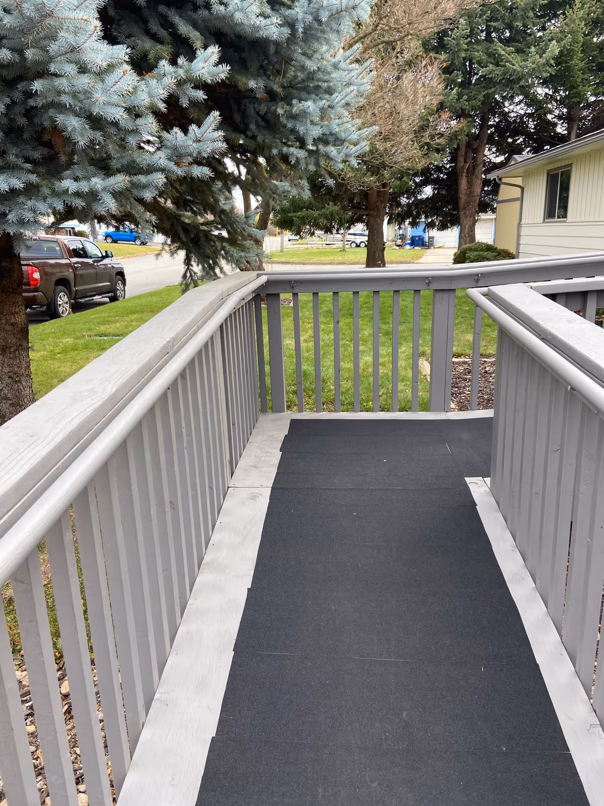 Exterior wooden ramp with gray railing beside a house and trees.