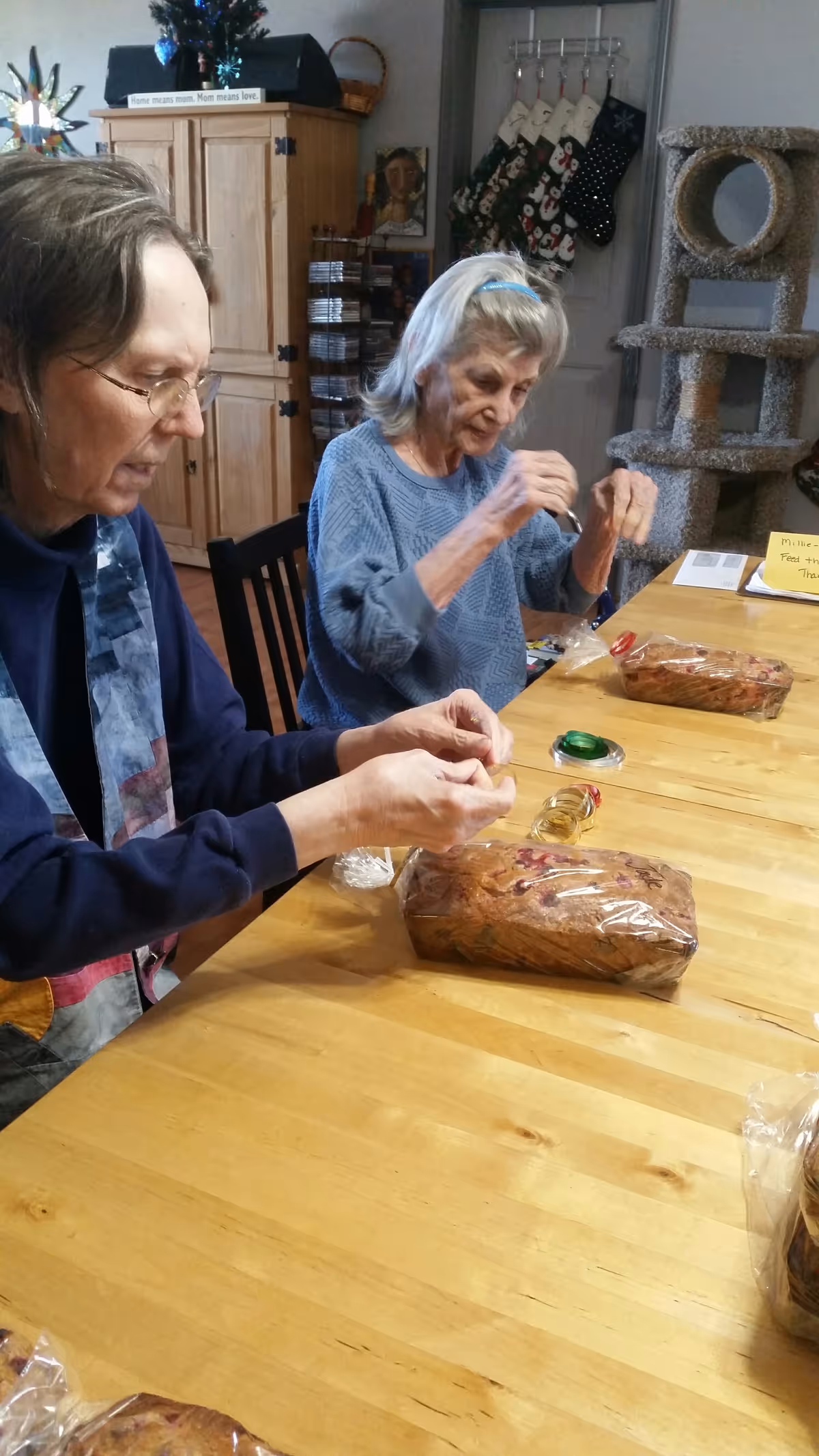 Two elderly women sitting at a wooden table, wrapping or unwrapping loaves of bread in plastic wrap. Behind them is a wooden cabinet, a cat tree, and a wall with hanging Christmas stockings.