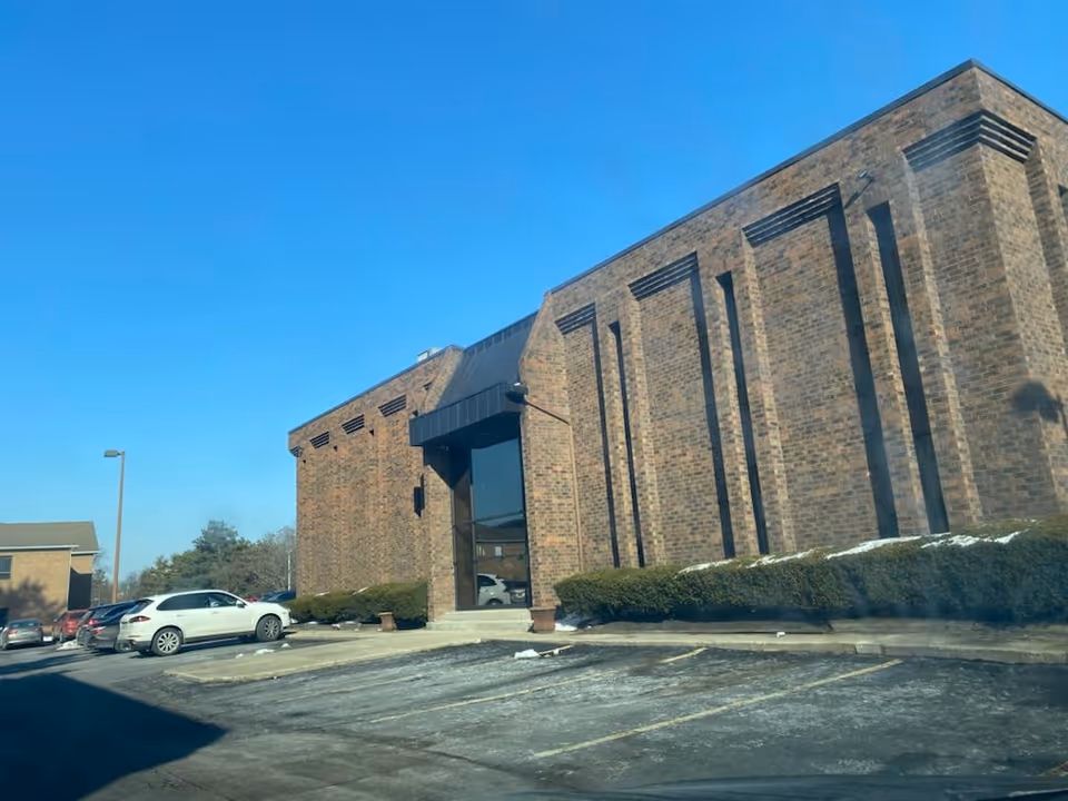 Brick building with tall facade, glass entrance, and lined parking lot under blue sky.