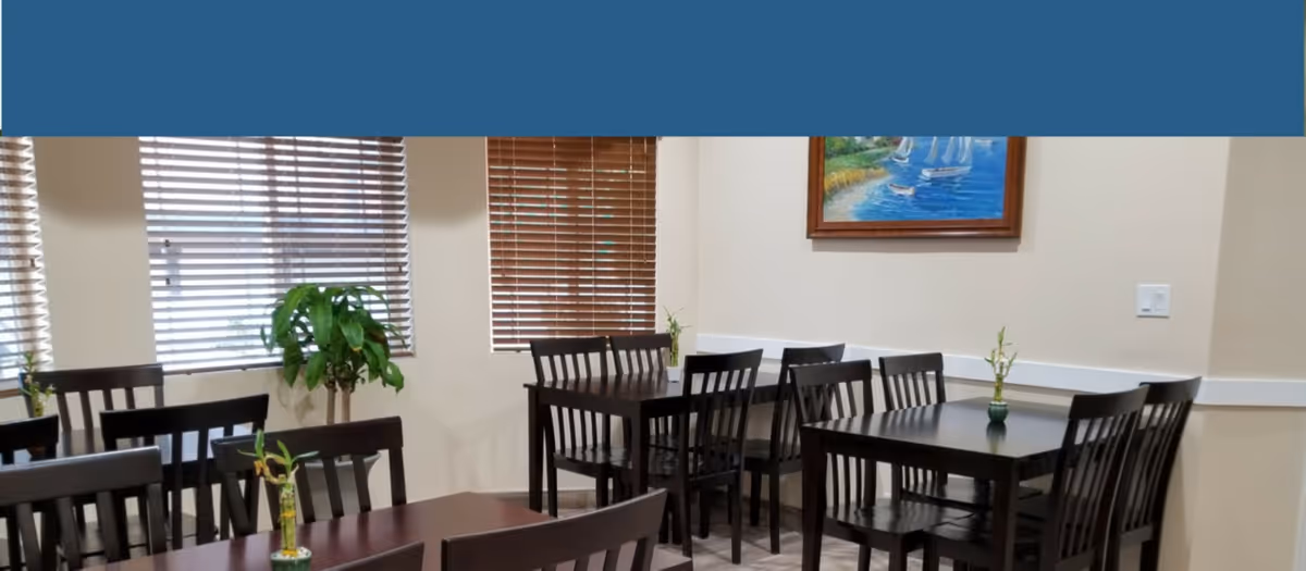 Dining room with dark wood tables and chairs, small vases and plants, window blinds and a framed painting on the wall.