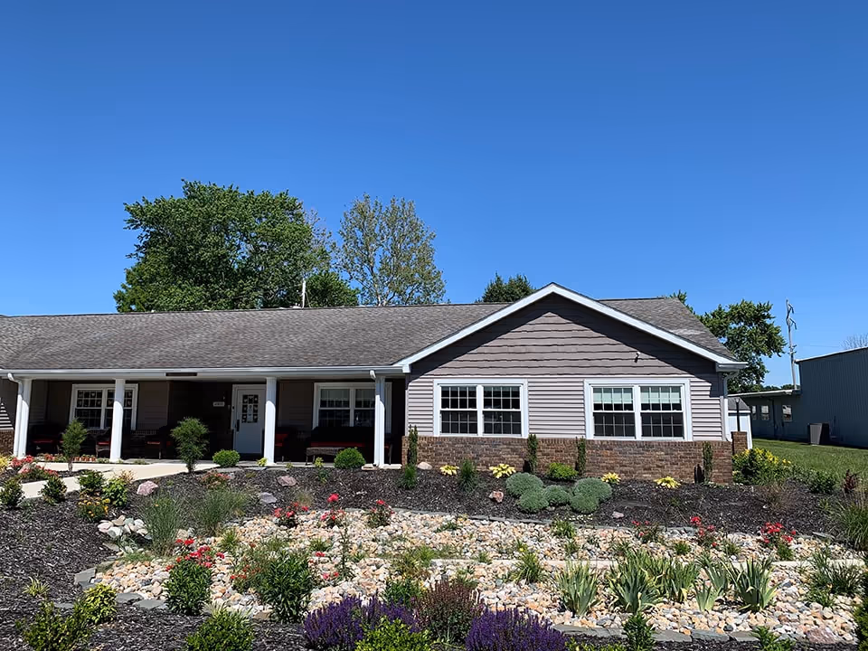 Single-story building with a gray roof and light purple siding, featuring a front porch with white columns and several windows. In front of the building is a landscaped garden with various plants, flowers, and decorative rocks under a clear blue sky.