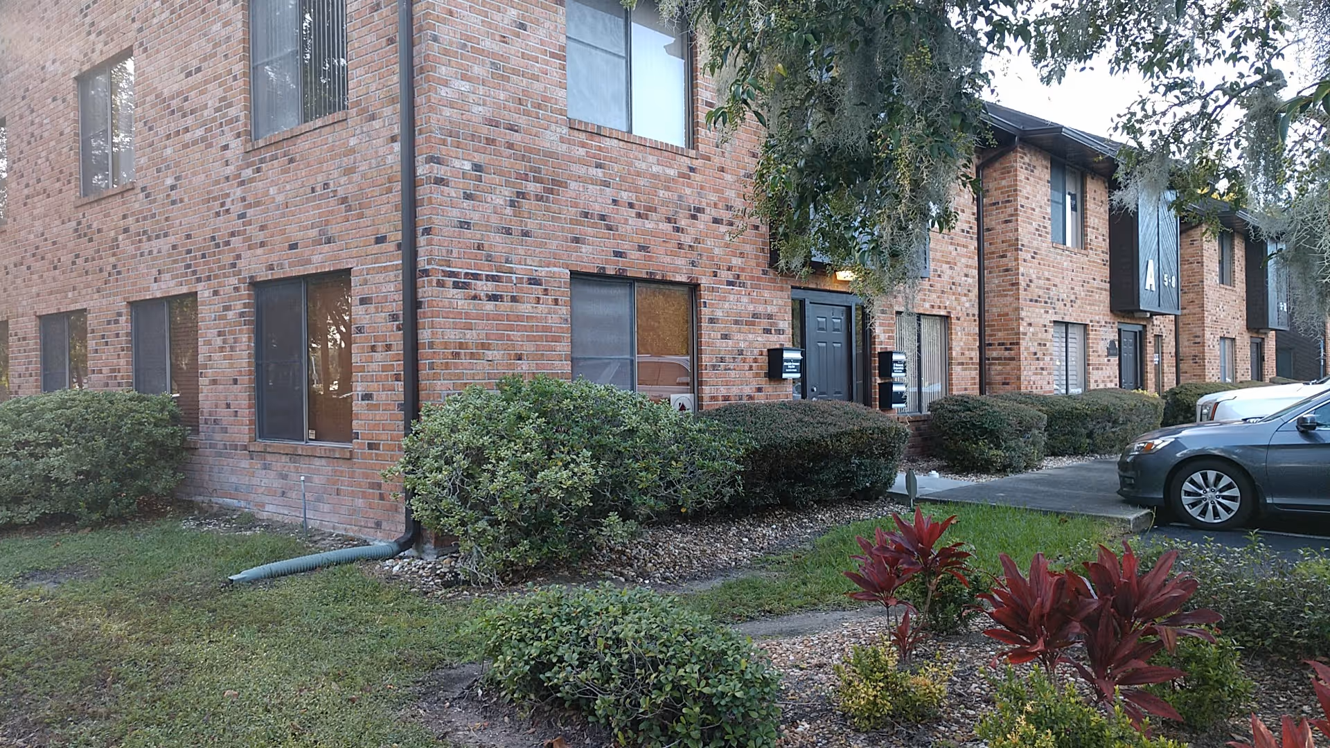 Brick senior living building exterior with manicured shrubs and parked cars along the front.
