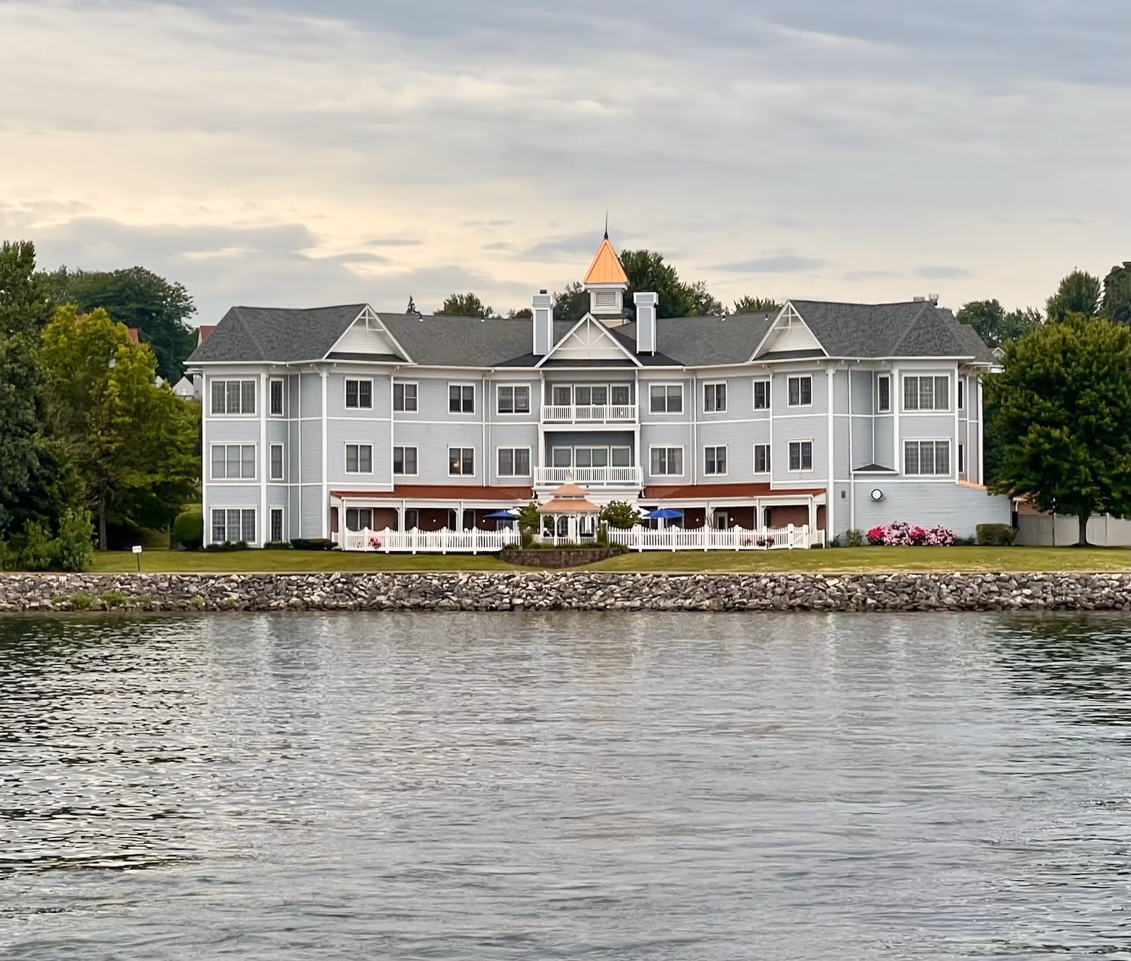A large, three-story residential building with gray siding and a peaked roof, situated beside a body of water with a rocky shoreline. The building features multiple windows, a central balcony, and a small gazebo in front, surrounded by a white fence and green lawn with trees on either side.