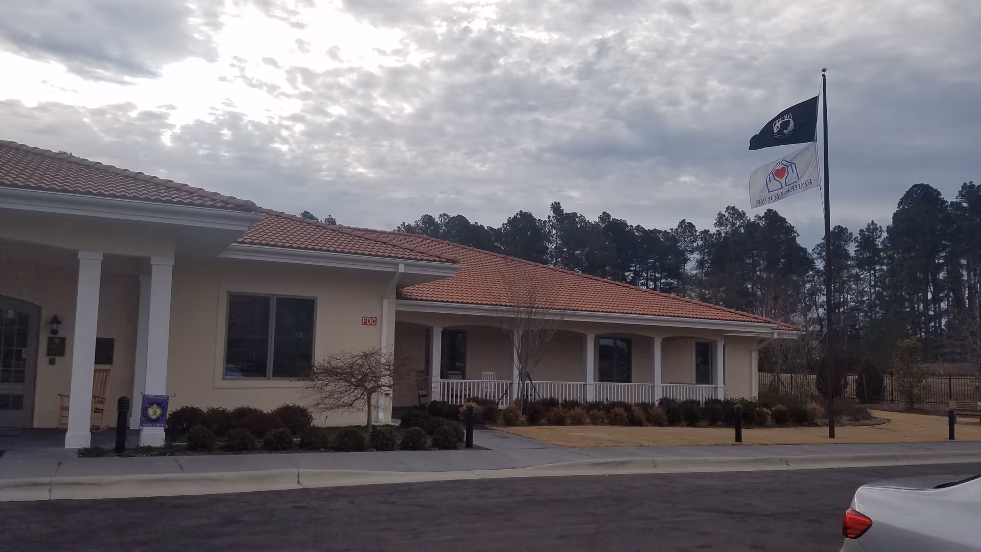 Single-story senior living facility with beige exterior, red-tiled roof, and flagpoles at the front.