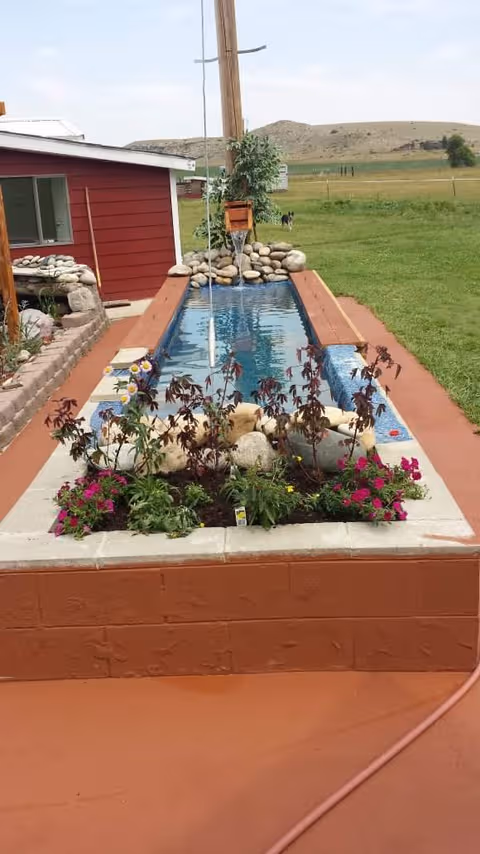 Outdoor water feature with a narrow pool, planter flowers, and a red building in rural setting.