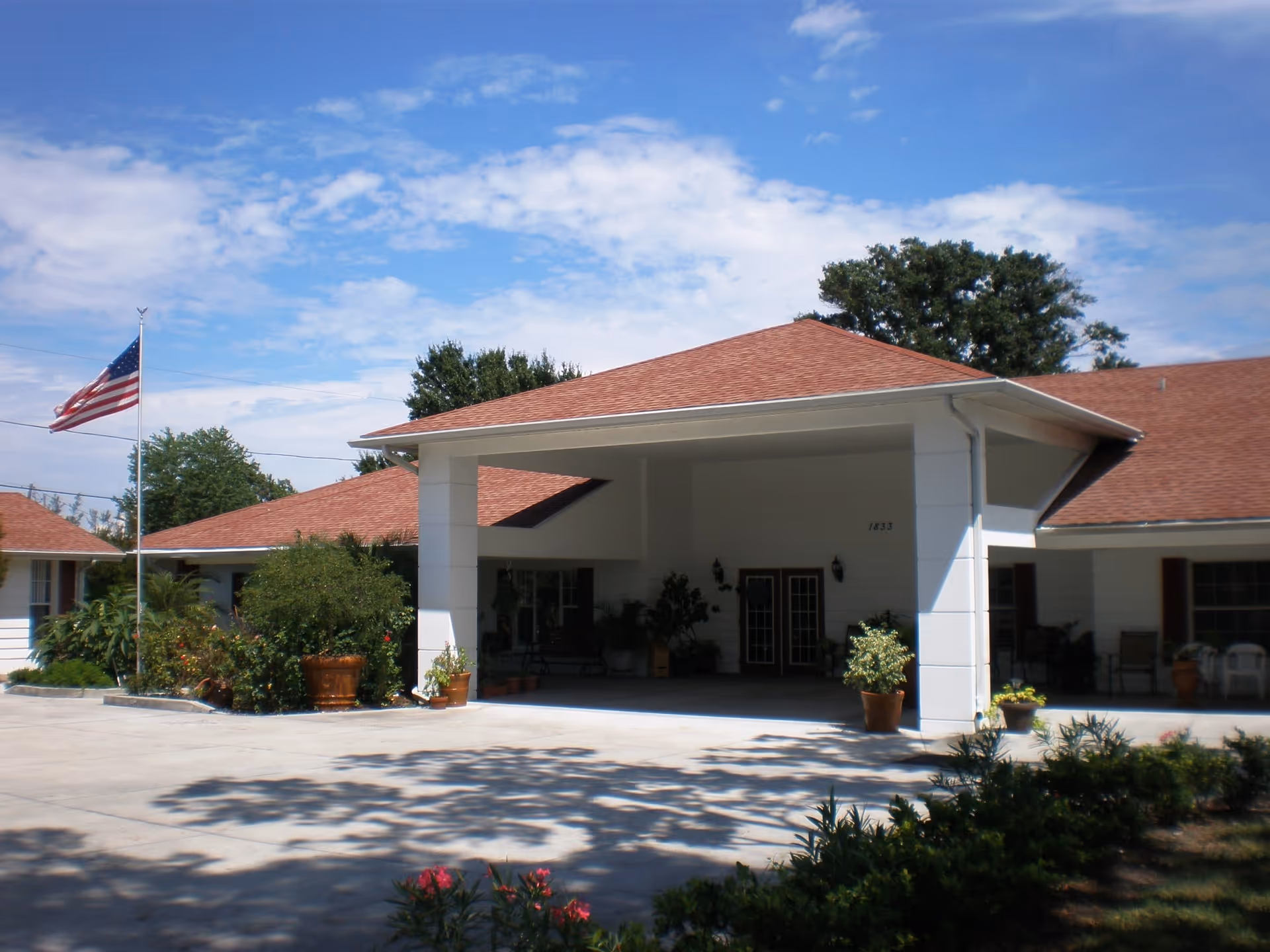 Exterior view of Royal Oaks Manor showing the entrance with a covered driveway, potted plants, an American flag on a flagpole, and a clear blue sky with some clouds.
